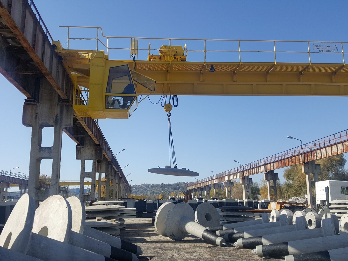 Żółty suwnicowy dźwig podnosi betonowy element na placu składowania prefabrykatów; w tle widoczne kolejne dźwigi i zaparkowana ciężarówka.