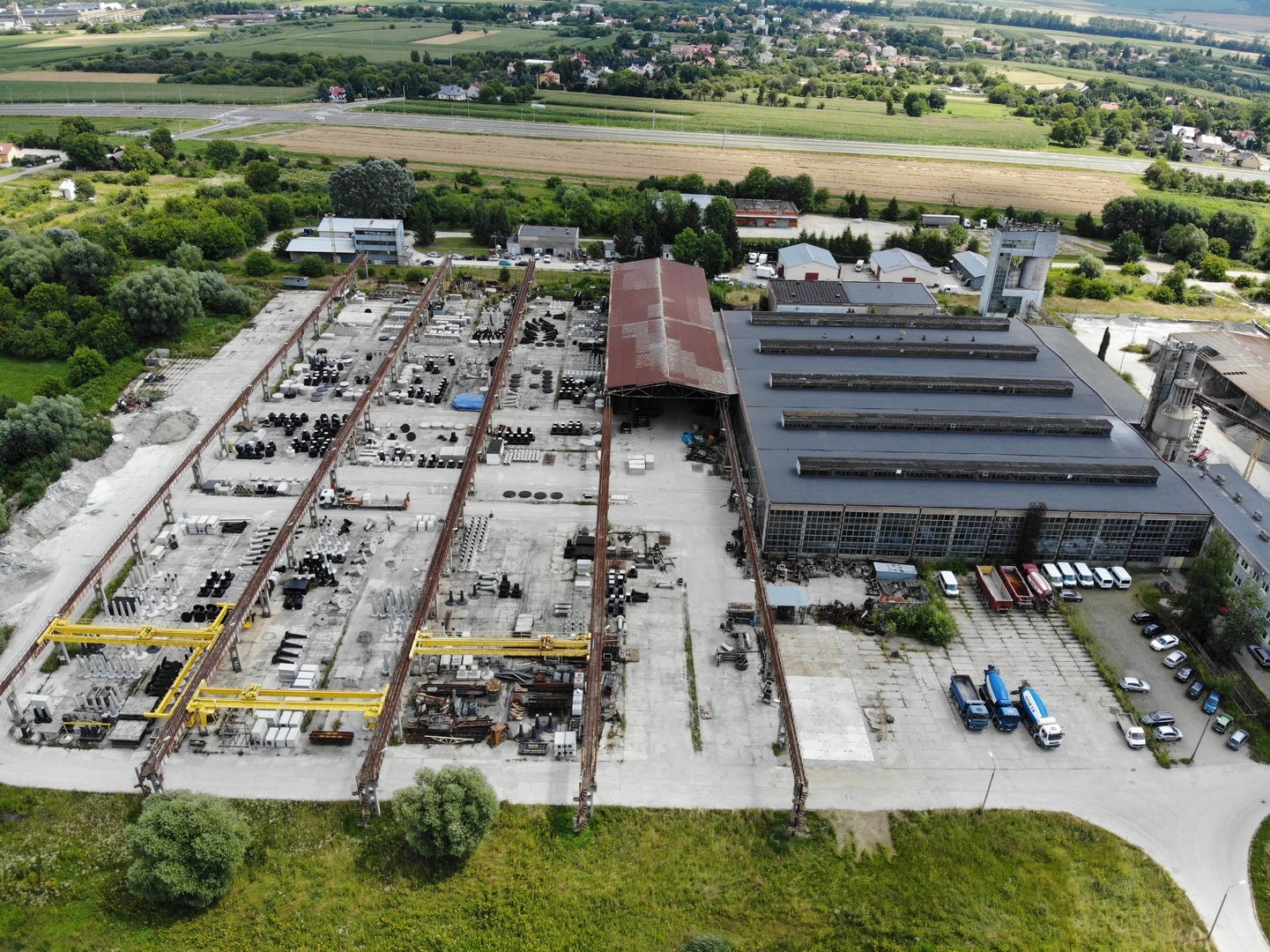 Widok z lotu ptaka na teren zakładu produkcyjnego z halami, suwnicami i składowanymi elementami betonowymi o różnych kształtach i rozmiarach. Na placu manewrowym zaparkowane betonomieszarki...