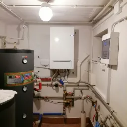 Kotłownia z zamontowanym zasobnikiem wody, piecem centralnego ogrzewania i rozbudowaną instalacją hydrauliczną z licznymi zaworami i rurami. Na zasobniku widoczna naklejka z logo firmy ERmet.