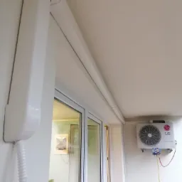 Biała jednostka zewnętrzna klimatyzacji LG z podłączonymi manometrami serwisowymi, widoczna z balkonu z oknem.