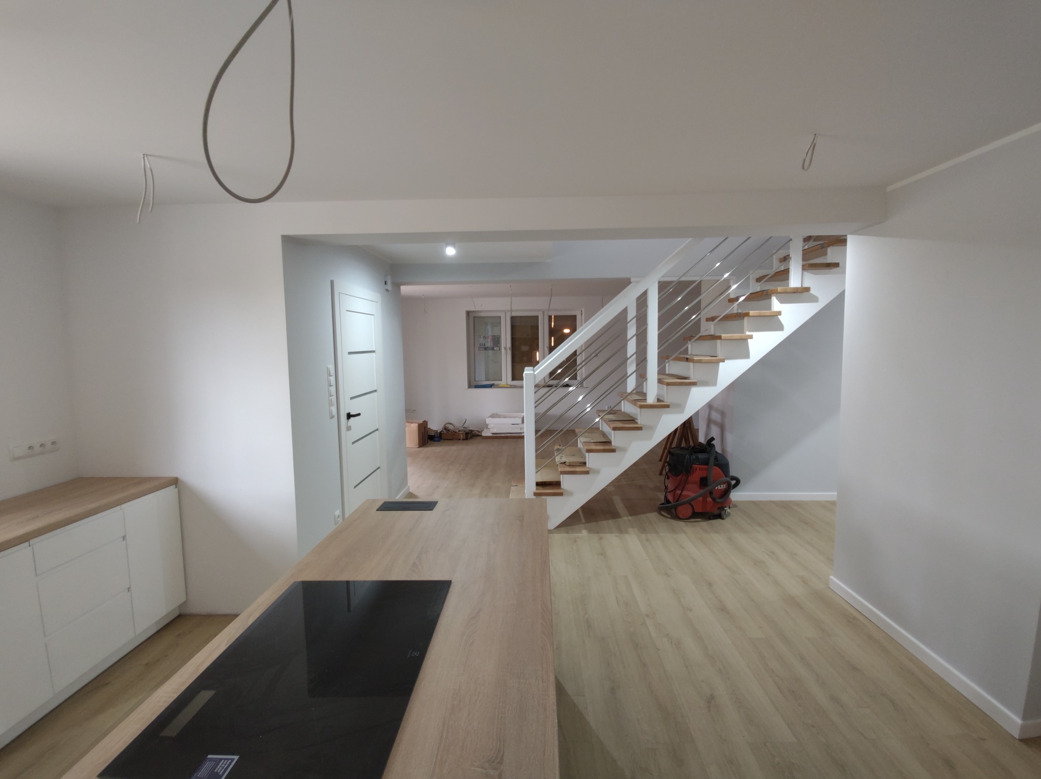 Wnętrze w trakcie prac wykończeniowych: widoczne schody z drewnianymi stopniami i metalową balustradą, zabudowa kuchenna z drewnianym blatem i płytą indukcyjną, oraz przewody elektryczne wystające...