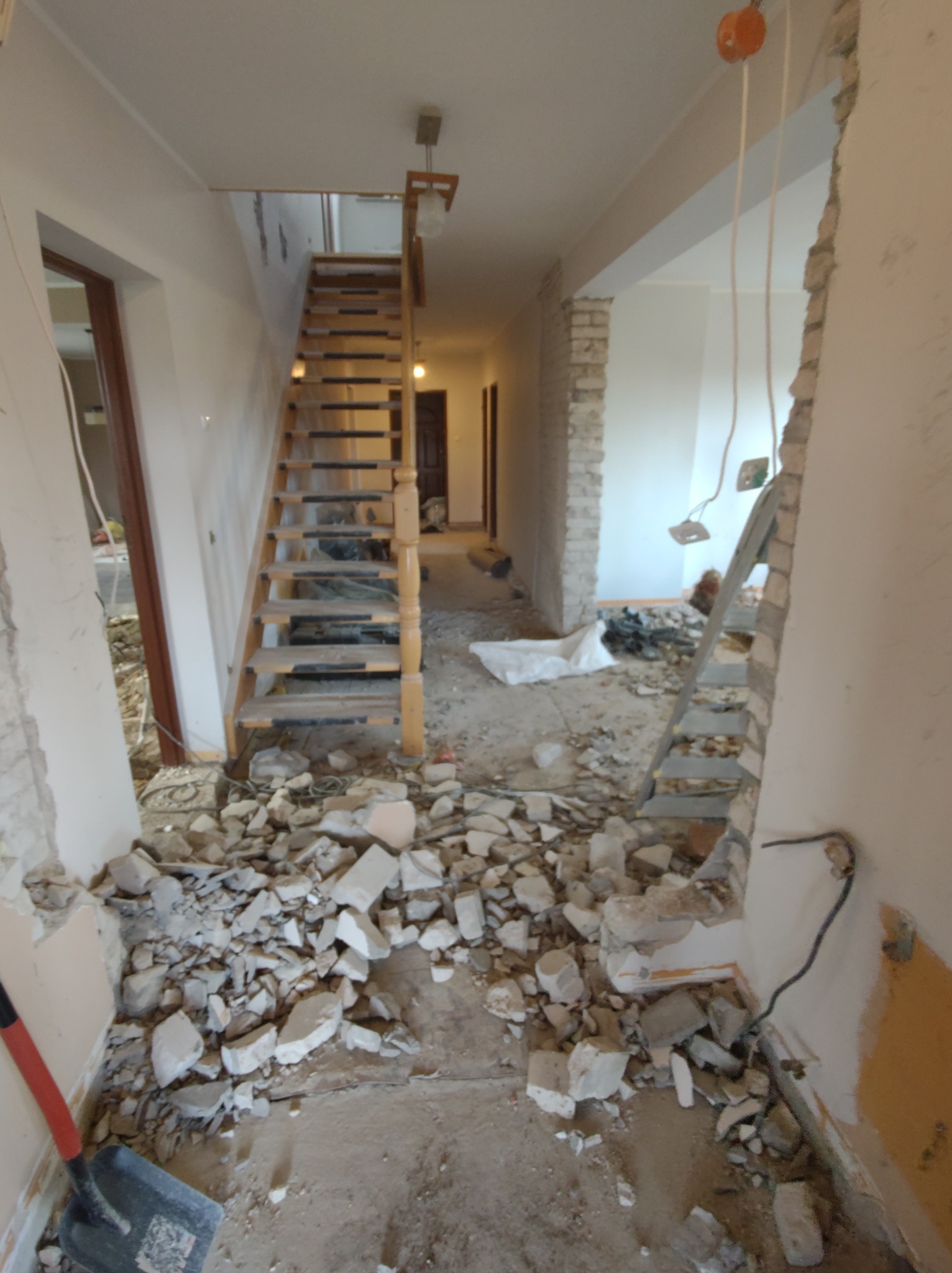 Korytarz w trakcie demontażu ścian działowych, widoczne gruz, schody drewniane, fragment instalacji elektrycznej i łopata.