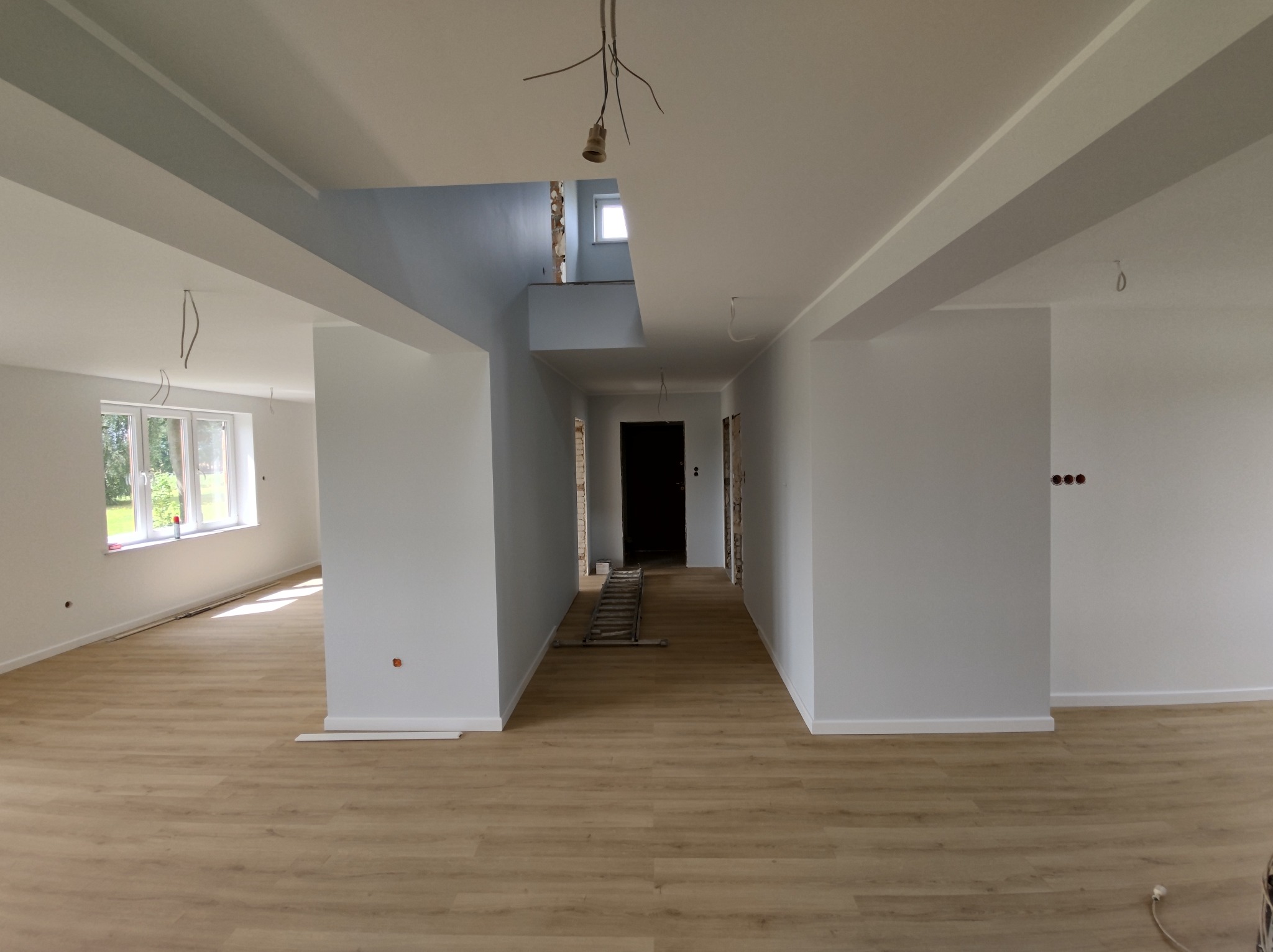 Przestronne wnętrze domu w trakcie prac wykończeniowych, z drewnopodobną podłogą, świeżo pomalowanymi na biało ścianami i widoczną klatką schodową z niebieskimi akcentami. Widoczne niedokończone...