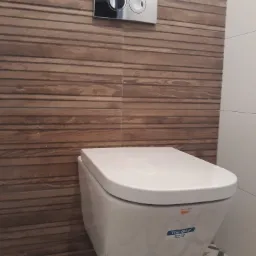 Wisząca toaleta Roca The Gap z chromowanym przyciskiem spłukującym na tle drewnopodobnych płytek ściennych i wzorzystej podłogi.