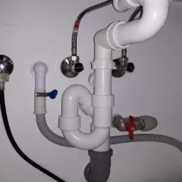 Kompletny system odpływowy pod zlewem z rur PCV, zaworami odcinającymi i elastycznymi wężami w białej szafce, widoczne połączenia i uszczelki.