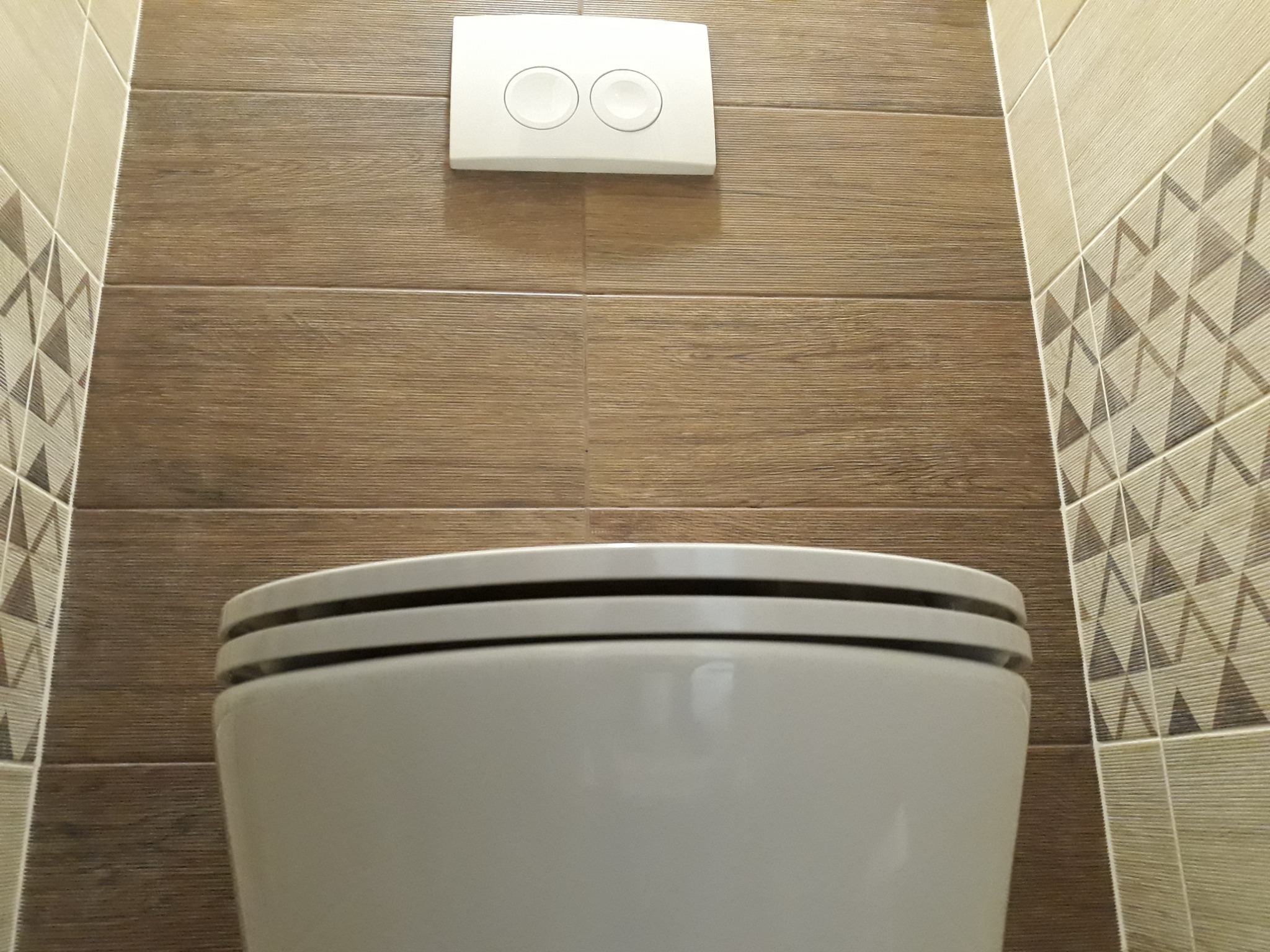 Nowoczesna toaleta wisząca z dwoma przyciskami spłukiwania na tle brązowych płytek imitujących drewno i dekoracyjnych płytek z geometrycznym wzorem.