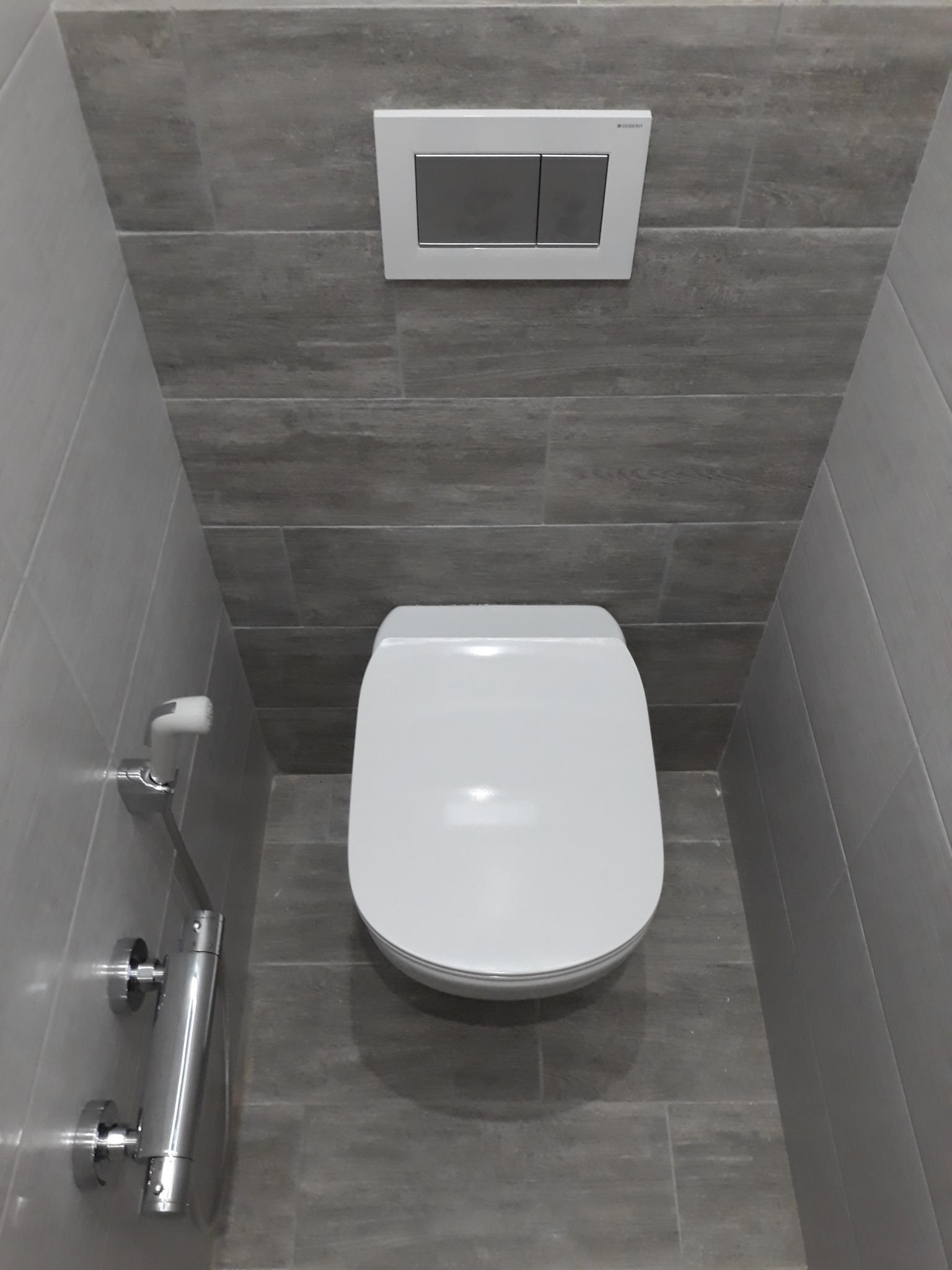 Nowoczesna toaleta podwieszana z szarymi płytkami imitującymi drewno i chromowaną słuchawką bidetową, widok z góry.