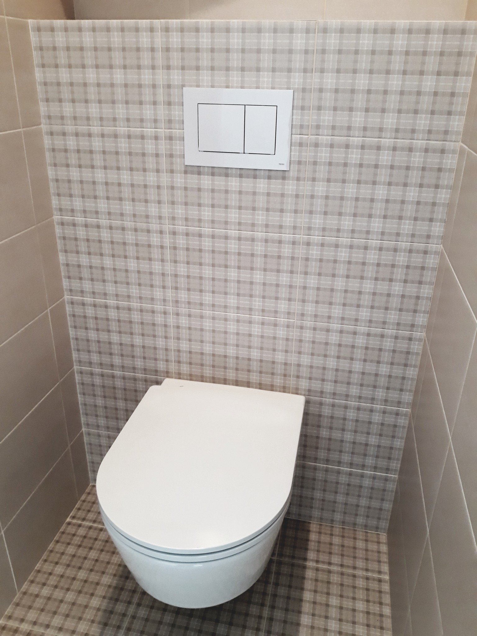 Zabudowana toaleta wisząca z przyciskiem spłukującym na ścianie wyłożonej kafelkami w kratę, z widocznymi fugami i kafelkami podłogowymi w tym samym wzorze.