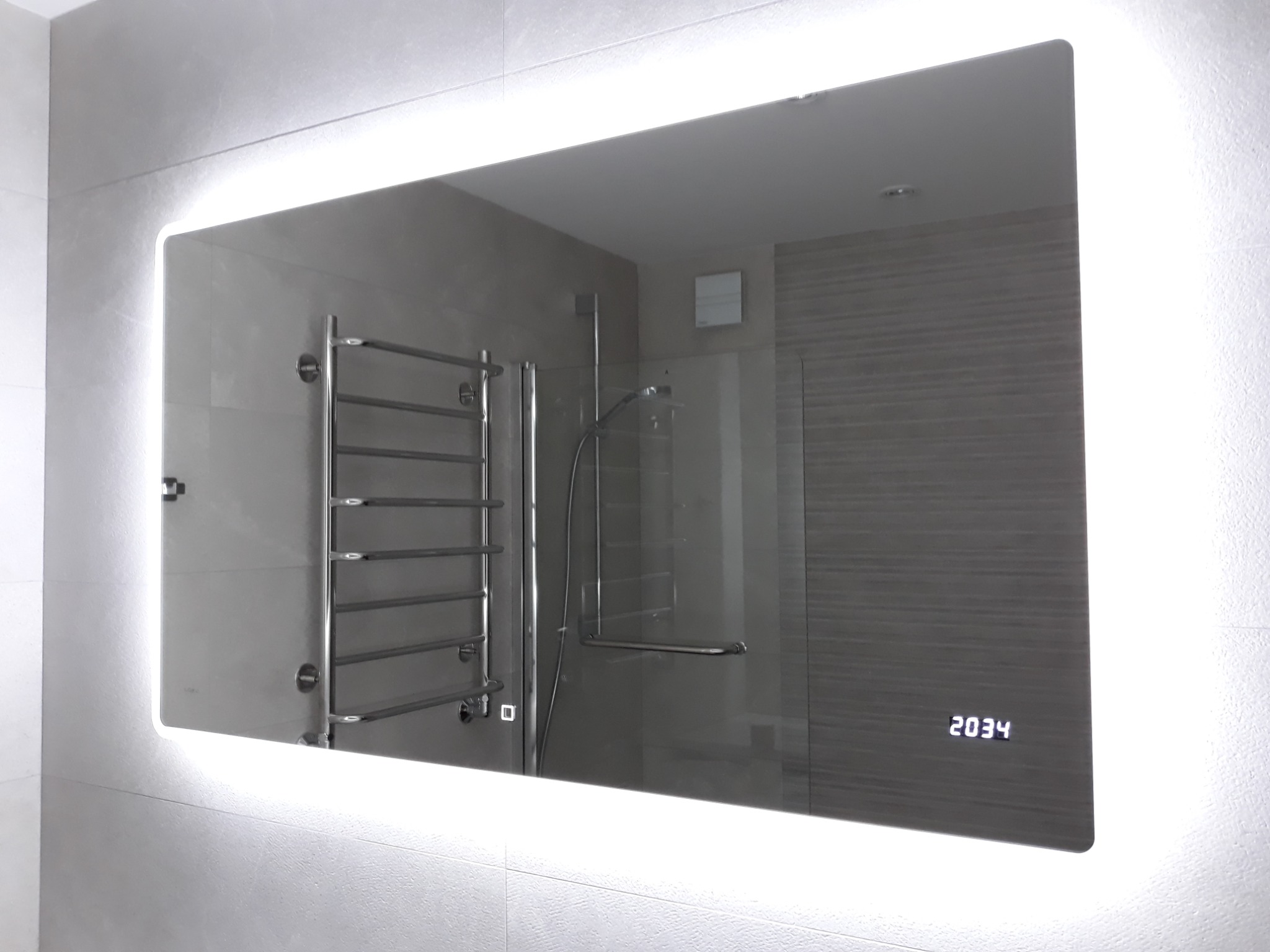 Podświetlane lustro łazienkowe z wyświetlaną godziną, odbijające grzejnik drabinkowy i kabinę prysznicową w nowoczesnej łazience.