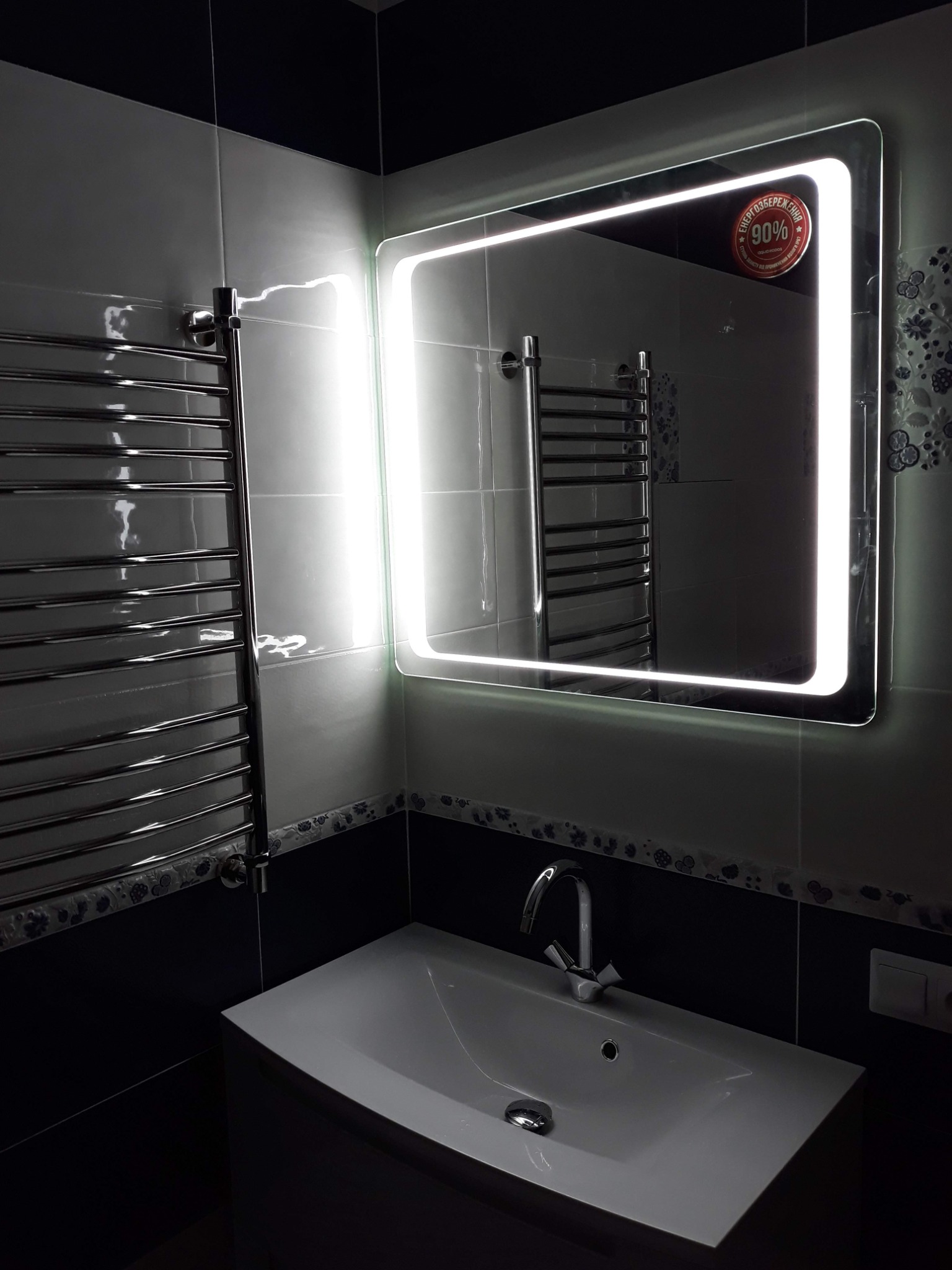 Nowoczesna łazienka z podświetlanym lustrem, grzejnikiem drabinkowym i umywalką nablatową, wykończona płytkami w odcieniach bieli i granatu z dekoracyjnym motywem roślinnym.