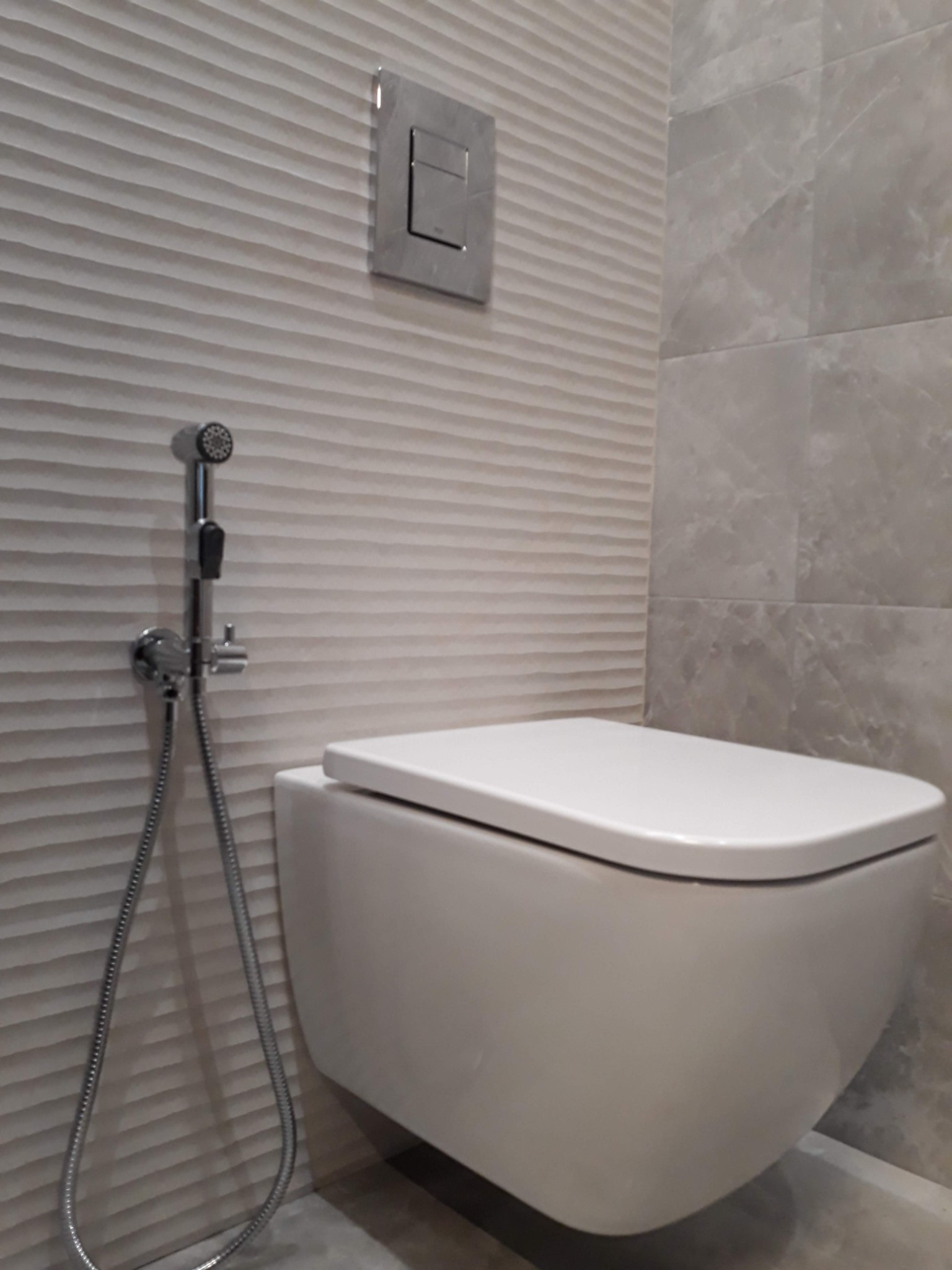 Nowoczesna toaleta podwieszana z bidetką, zamontowana na ścianie z dekoracyjnymi, poziomymi żłobieniami, obok płytek imitujących marmur.