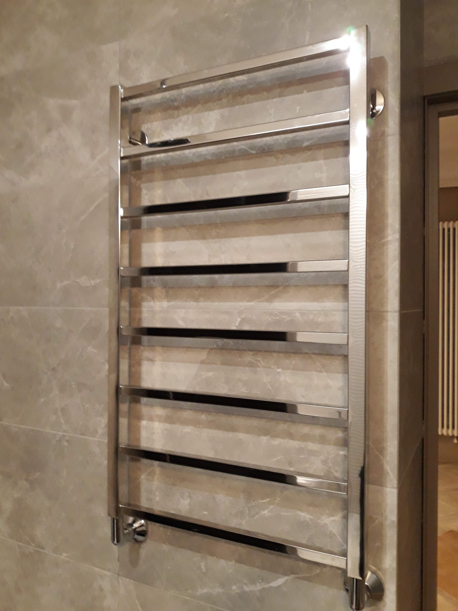 Chromowany grzejnik łazienkowy drabinkowy, zamontowany na ścianie z marmurowych płytek, widok z boku, z widocznym fragmentem grzejnika pokojowego w tle.