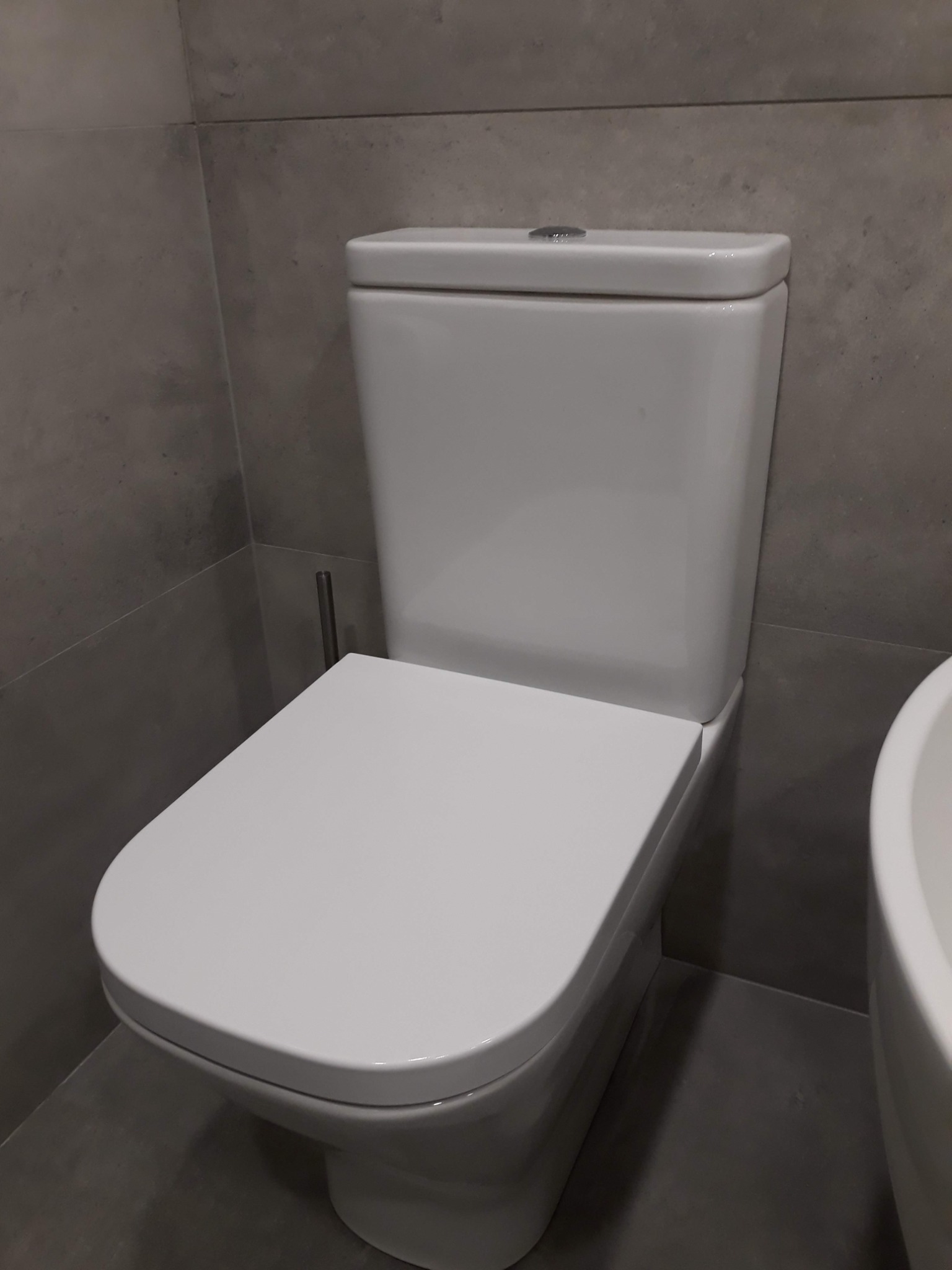 Zamontowana, biała toaleta kompaktowa z ceramiczną spłuczką i deską sedesową, widoczna częściowo umywalka, szare płytki na ścianach i podłodze.