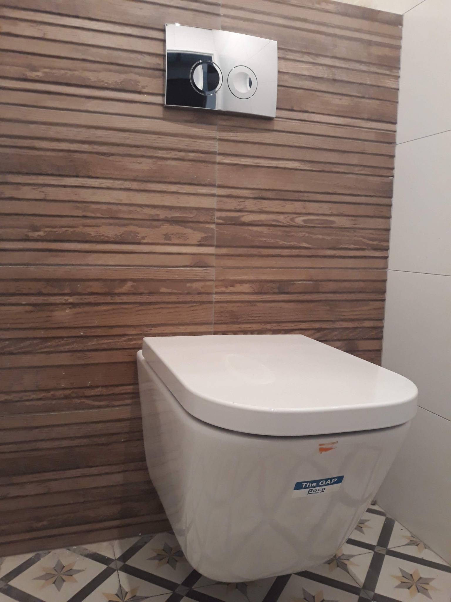 Wisząca toaleta Roca The Gap z chromowanym przyciskiem spłukującym na tle drewnopodobnych płytek ściennych i wzorzystej podłogi.