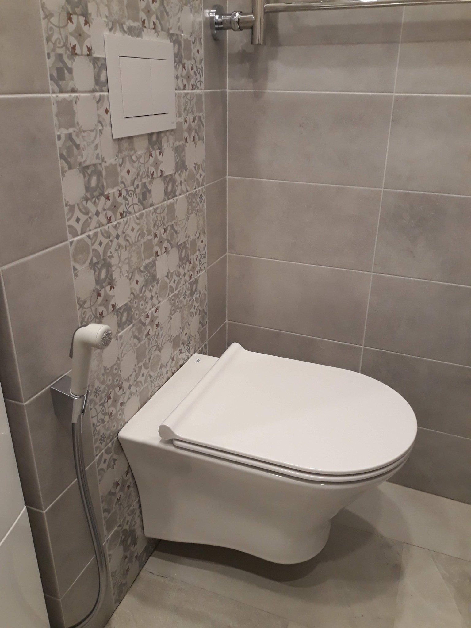 Nowoczesna, podwieszana toaleta z bidetką w łazience wyłożonej szarymi płytkami i dekoracyjnym pasem płytek wzorzystych.