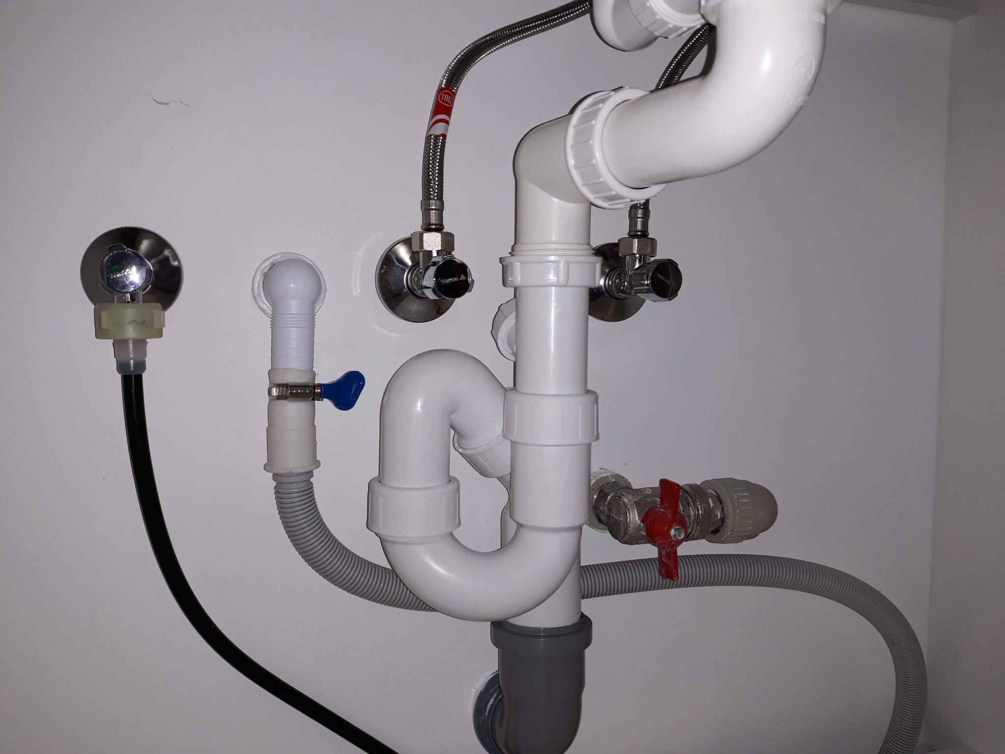 Kompletny system odpływowy pod zlewem z rur PCV, zaworami odcinającymi i elastycznymi wężami w białej szafce, widoczne połączenia i uszczelki.