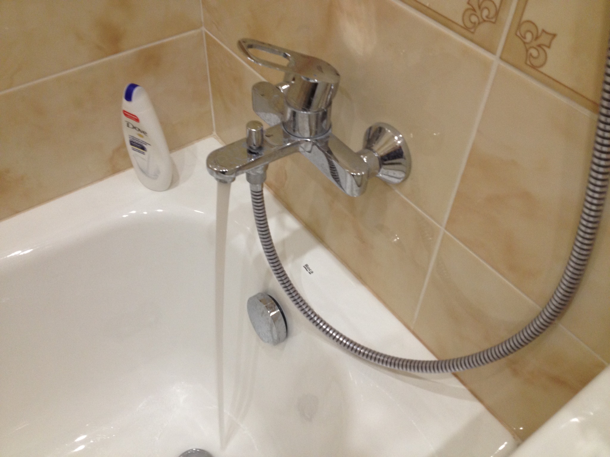 Woda leje się z chromowanej baterii wannowej w łazience z beżowymi płytkami, obok butelka kosmetyku