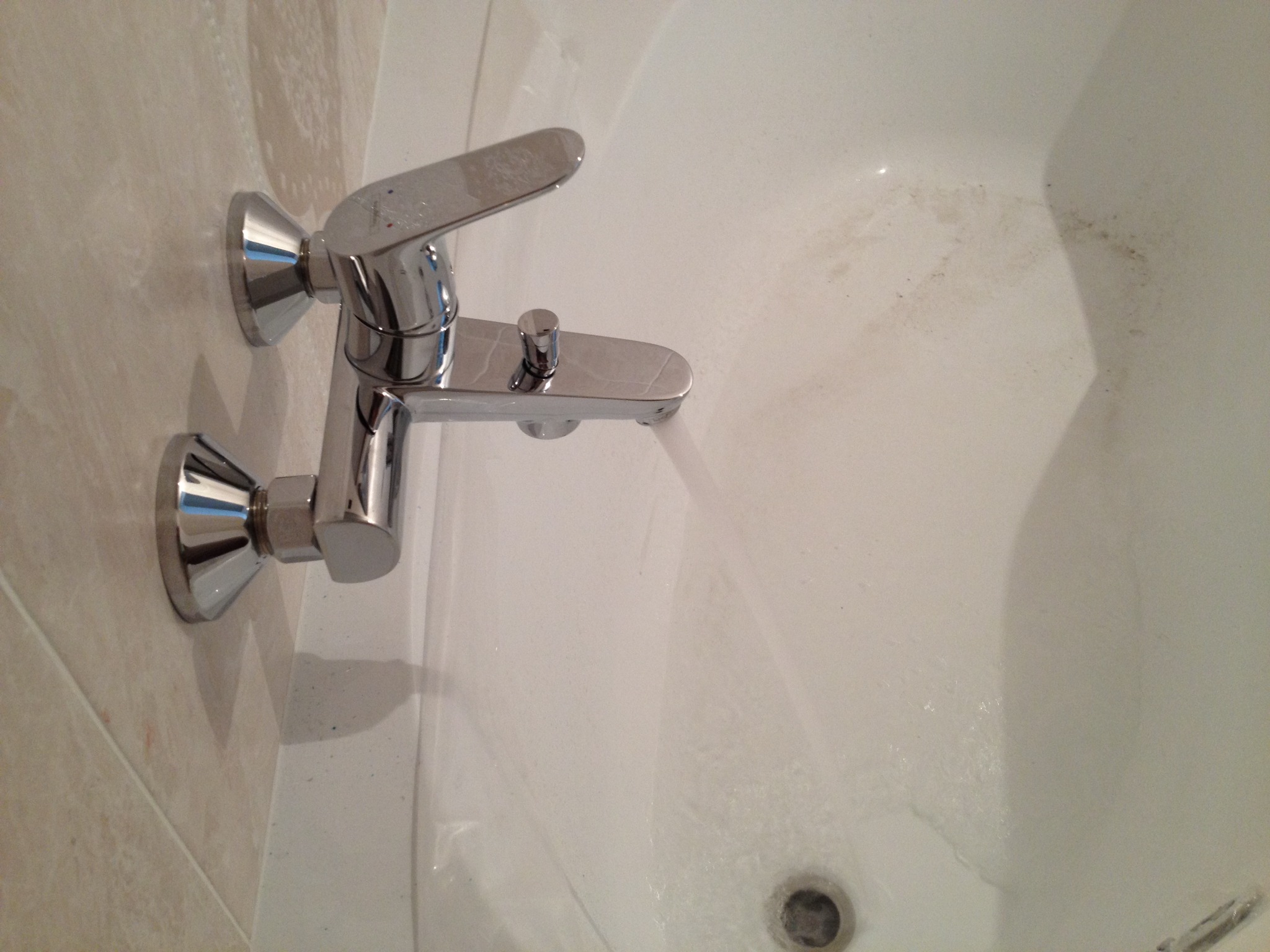 Bateria wannowa chromowana, zamontowana na jasnej ścianie z płytek, widoczna część wanny z odpływem.