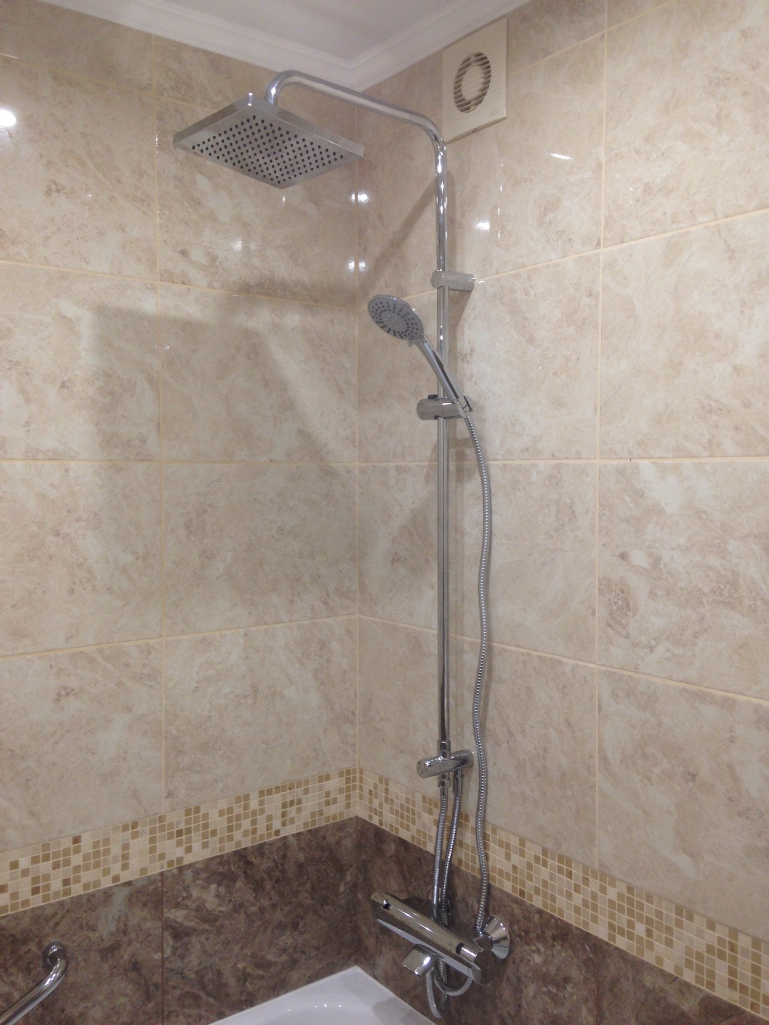 Nowoczesny panel prysznicowy z deszczownicą i słuchawką, zamontowany w kabinie wyłożonej beżowymi płytkami imitującymi marmur i ciemnobrązową mozaiką.