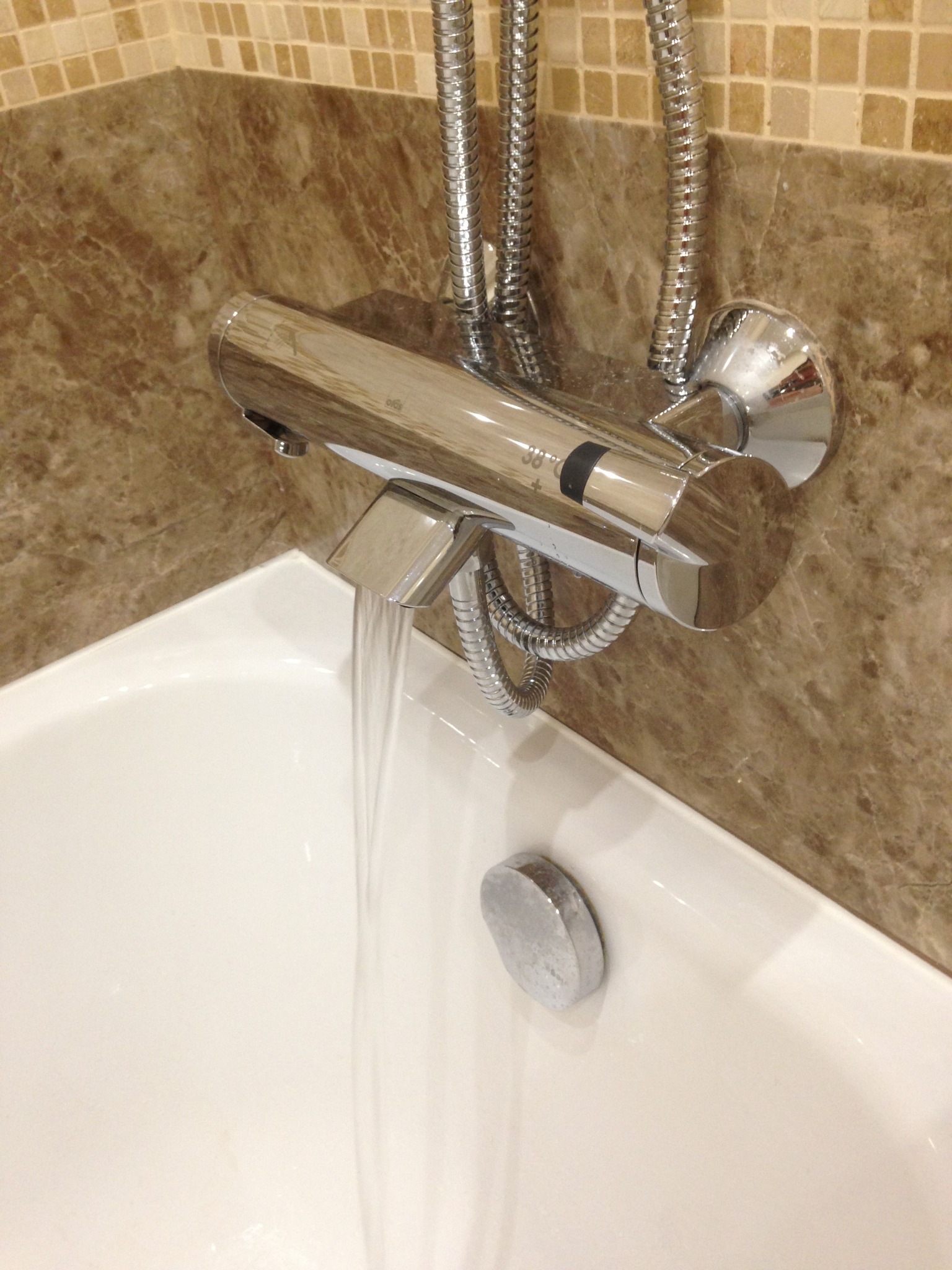 Bateria wannowa termostatyczna chromowana zamontowana na marmurowej ścianie, woda leje się do białej wanny.