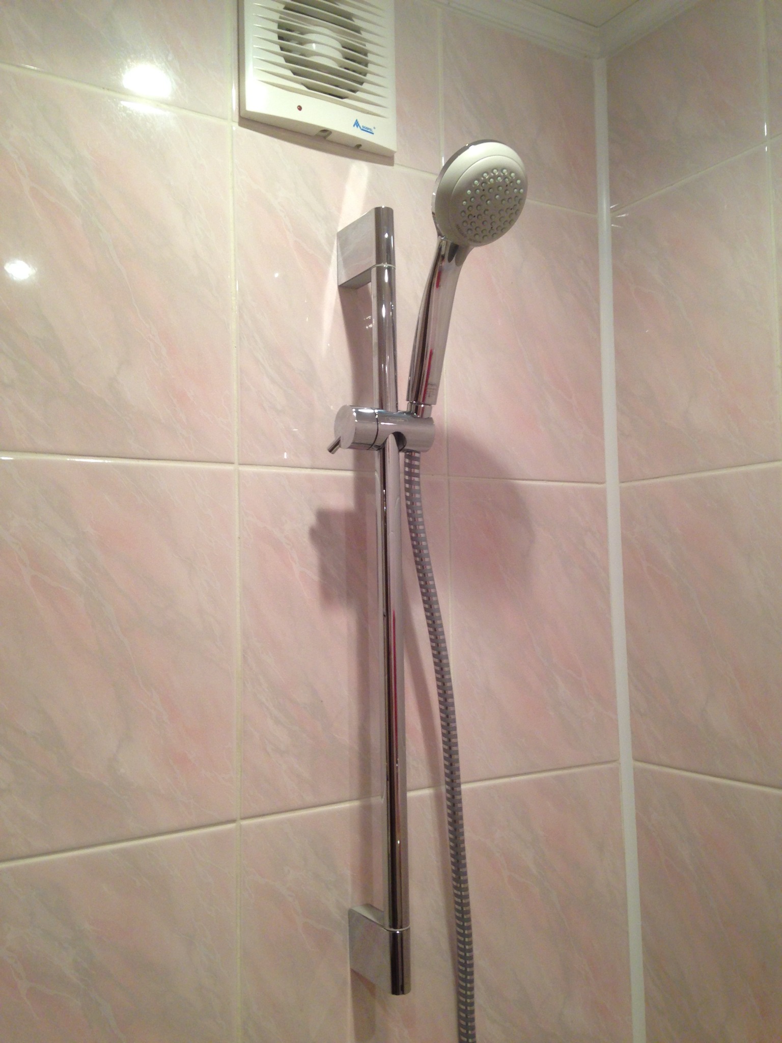 Chromowana słuchawka prysznicowa z regulowanym drążkiem zamontowana na ścianie z różowych płytek ceramicznych w łazience, widoczny wentylator w górnym rogu.