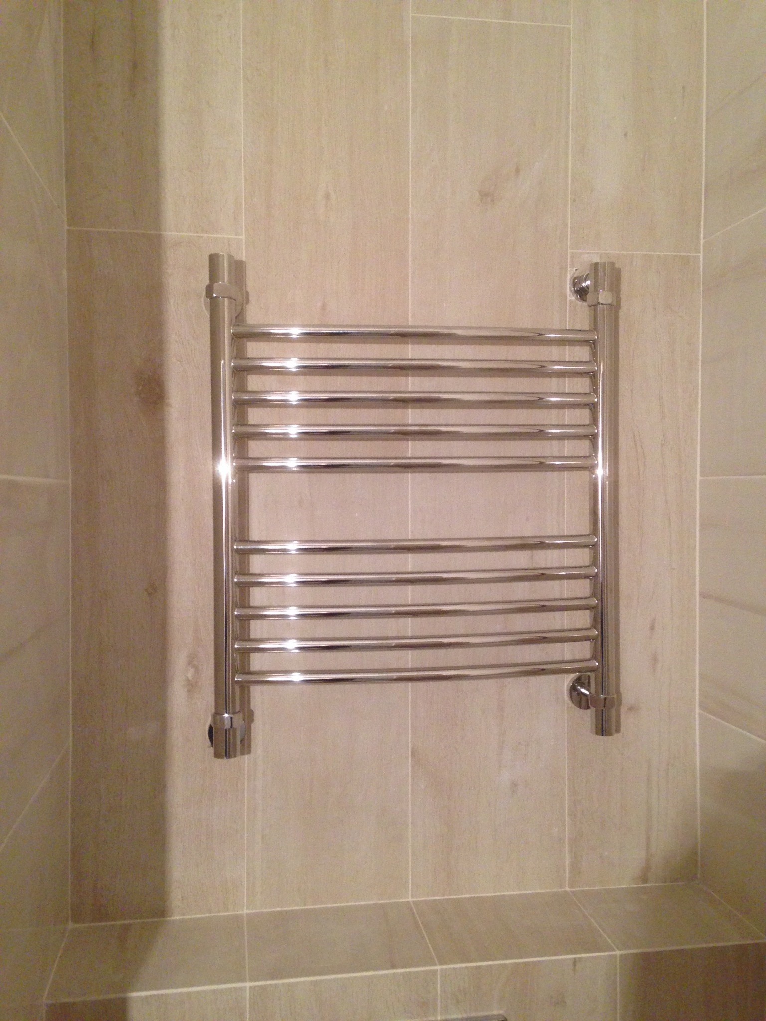 Chromowany, drabinkowy grzejnik łazienkowy zamontowany na beżowych, pionowych płytkach ceramicznych.