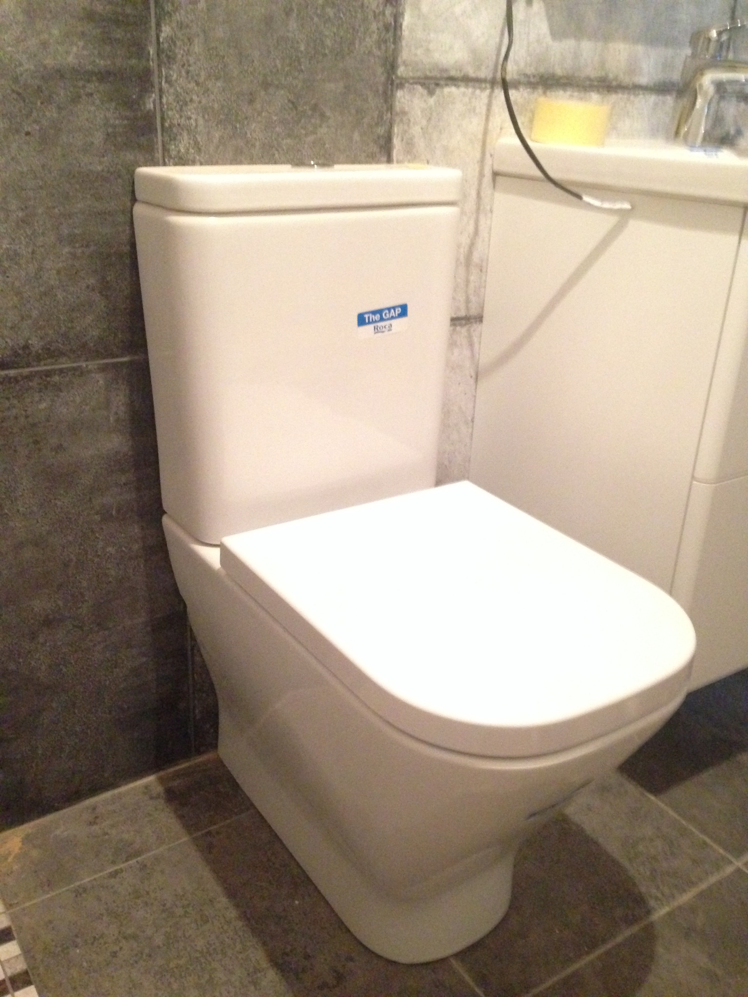 Biała, kompaktowa toaleta z zamontowaną deską sedesową, model The Gap firmy Roca, na tle szarych płytek ściennych i podłogowych, obok biała szafka umywalkowa.