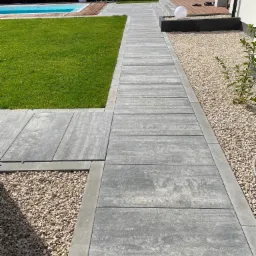 Szara ścieżka z prostokątnych płyt betonowych prowadzi przez trawnik i żwirowe rabaty w kierunku basenu i tarasu.