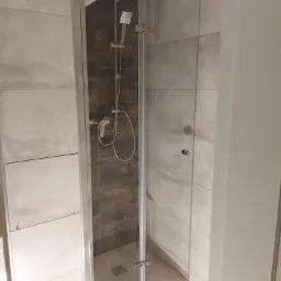 Kabina prysznicowa z przeszklonymi drzwiami i panelami, chromowana armatura, płytki w odcieniach beżu i brązu, widoczny odpływ liniowy.