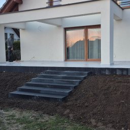 Tomasz Kuczmik Terra-Bruk brukarstwo i usługi ziemne - Nowoczesny taras z kamiennymi schodami i kostką, świeżo obsypany ziemią. Widoczny fragment domu z oknem i zadaszeniem. Prace wykończeniowe w toku.