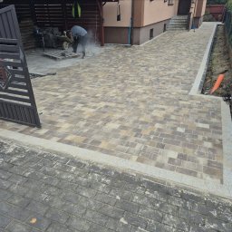 Tomasz Kuczmik Terra-Bruk brukarstwo i usługi ziemne - Ułożona kostka brukowa w odcieniach beżu i szarości tworzy wzór na podjeździe. W tle pracownik docina kostkę, widoczne ogrodzenie panelowe i brama wjazdowa.