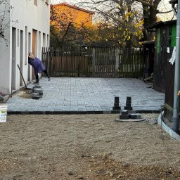 Tomasz Kuczmik Terra-Bruk brukarstwo i usługi ziemne - Fragment układania szarej kostki brukowej na podsypce żwirowej, widoczne narzędzia i częściowo ułożona nawierzchnia przy budynku z drewnianym ogrodzeniem w tle.