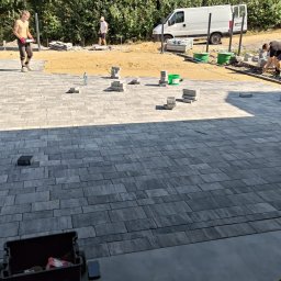 Tomasz Kuczmik Terra-Bruk brukarstwo i usługi ziemne - Układanie szarej kostki brukowej na posesji. Widoczni pracownicy, przygotowane materiały i częściowo ułożona nawierzchnia. Praca w toku, jasne oświetlenie.