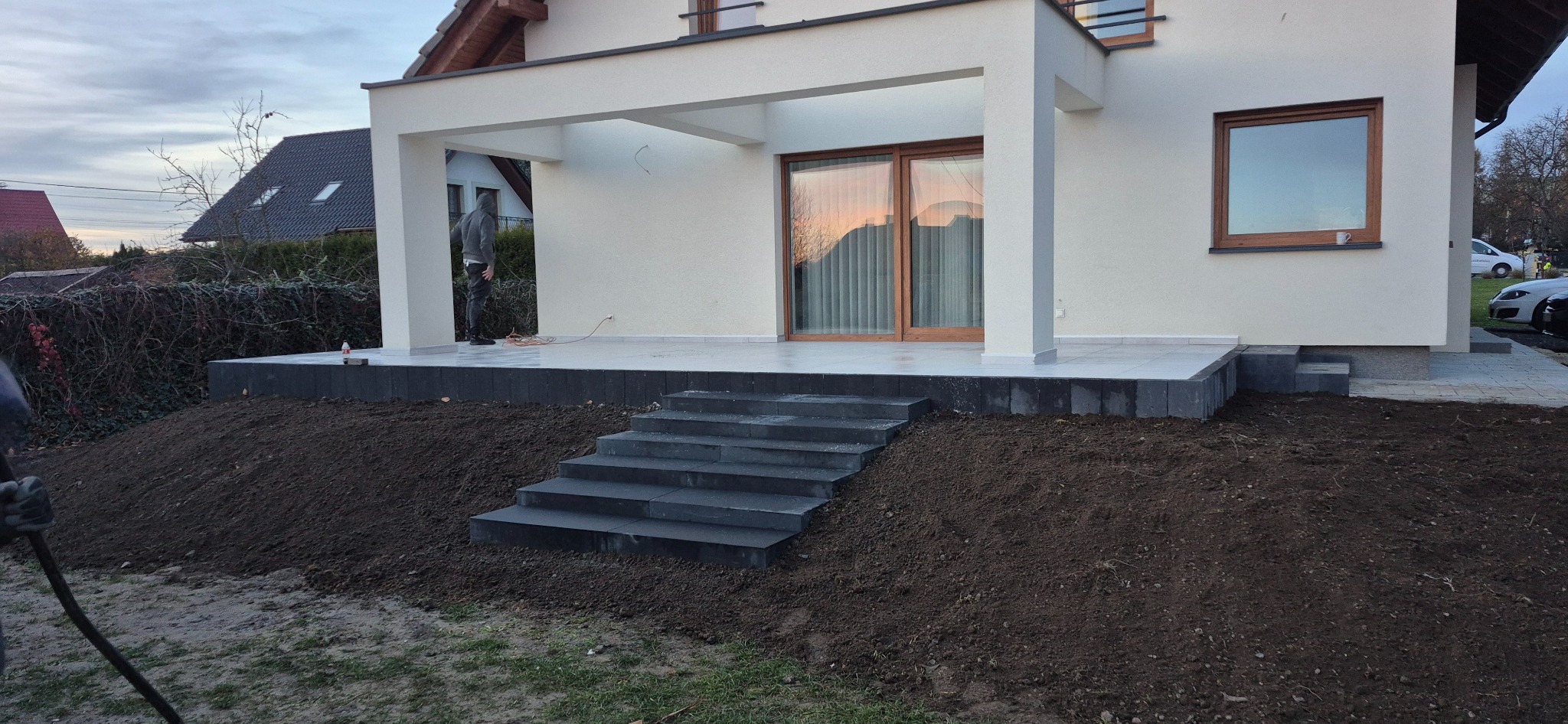 Nowoczesny taras z kamiennymi schodami i kostką, świeżo obsypany ziemią. Widoczny fragment domu z oknem i zadaszeniem. Prace wykończeniowe w toku.