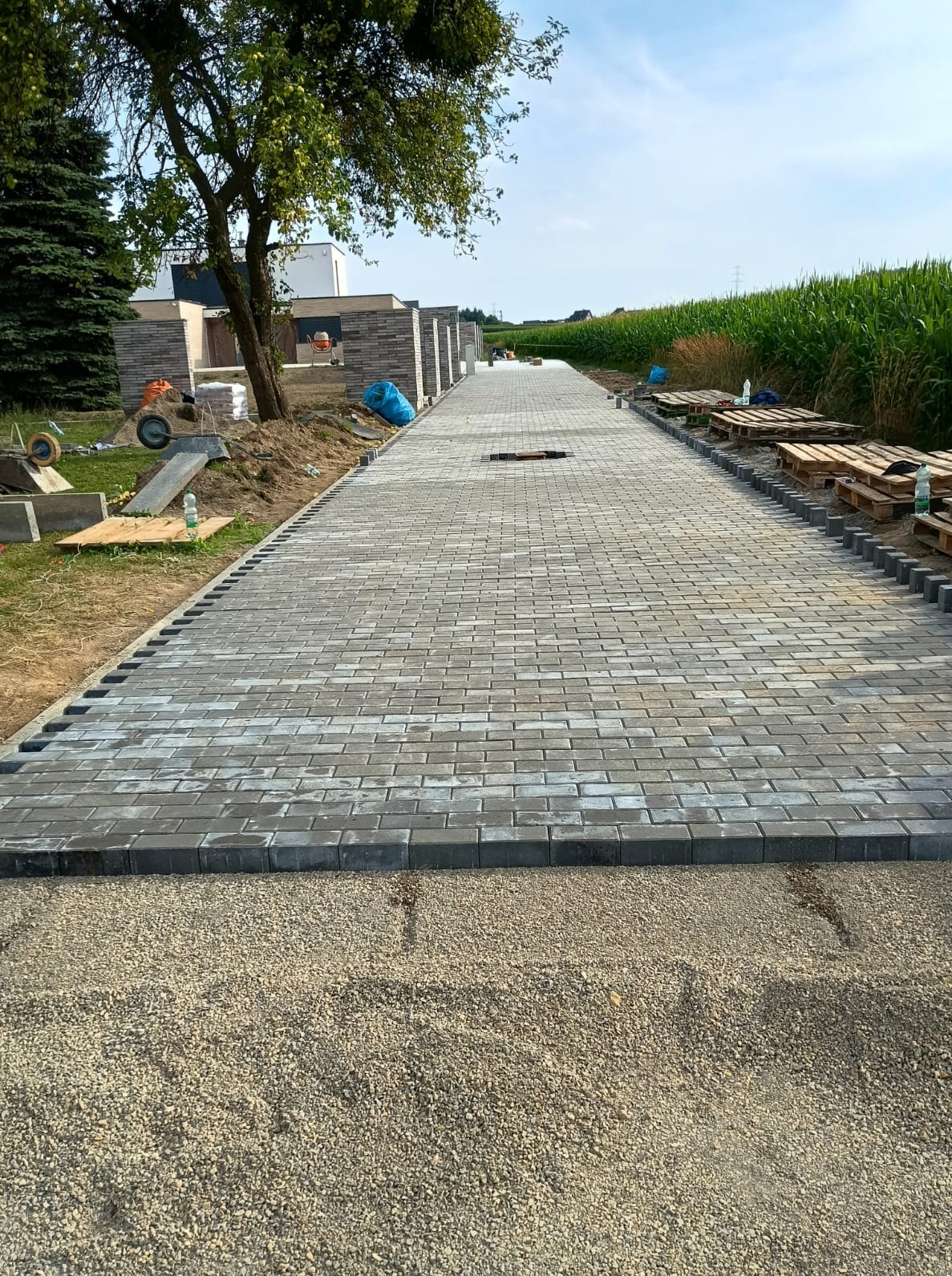 Długa, szara ścieżka z kostki brukowej prowadząca wzdłuż nowoczesnego domu, z widocznym podłożem z kruszywa i paletami w tle, w słoneczny dzień.