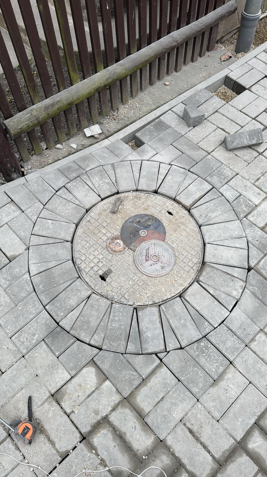 Układanie kostki brukowej wokół studzienki kanalizacyjnej. Widoczne tarcze do cięcia kostki i miarka. Kostka ułożona w wzór jodełki.