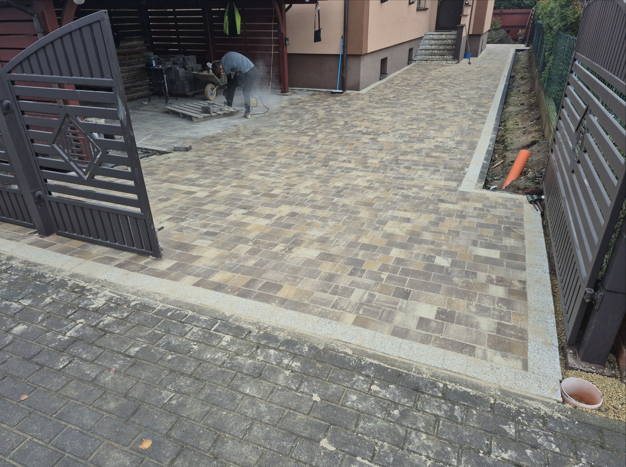 Ułożona kostka brukowa w odcieniach beżu i szarości tworzy wzór na podjeździe. W tle pracownik docina kostkę, widoczne ogrodzenie panelowe i brama wjazdowa.