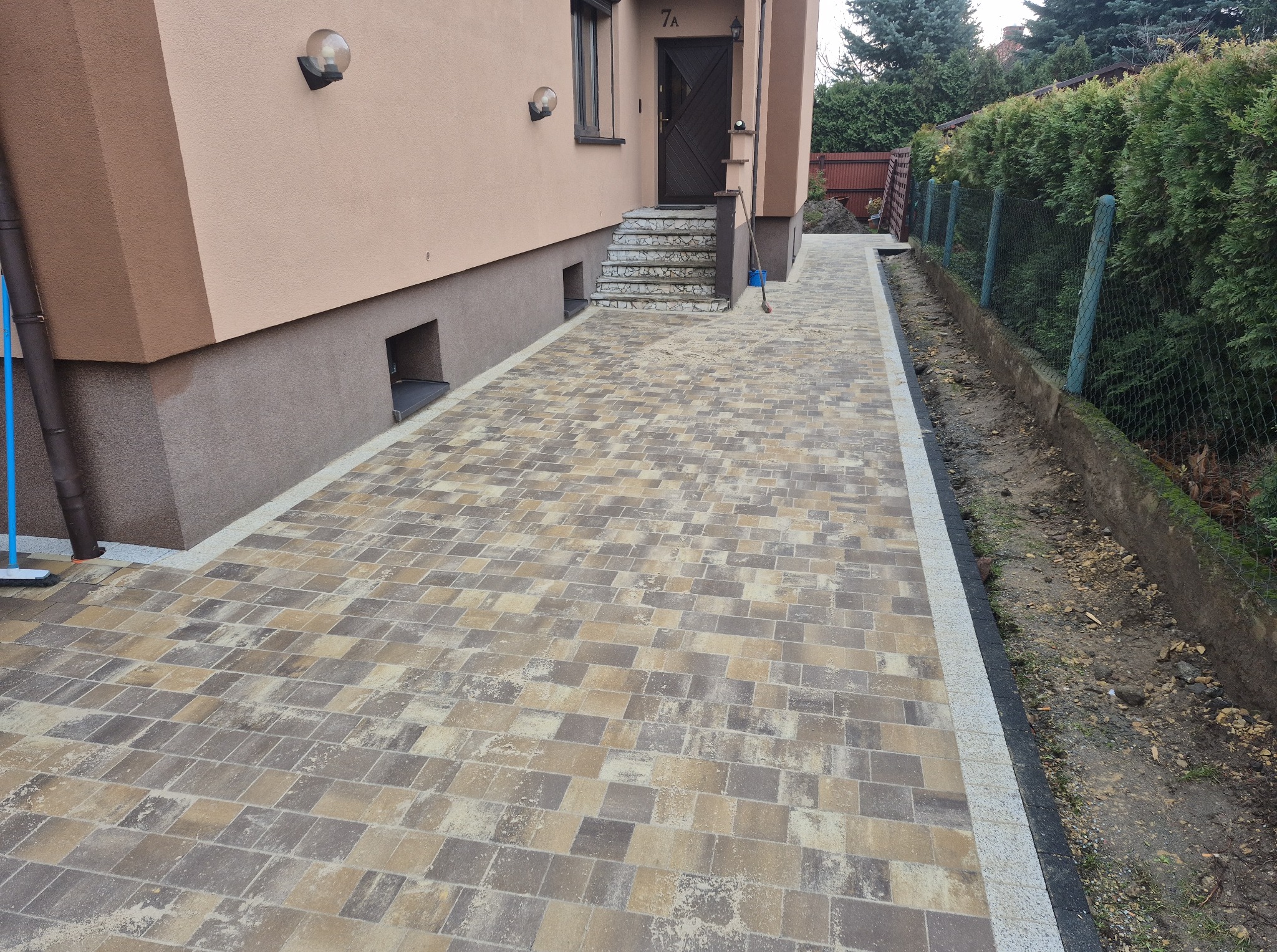 Ułożona kostka brukowa w odcieniach beżu i szarości tworzy ścieżkę obok domu z numerem 7A, prowadzącą do schodów wejściowych. Widoczny fragment ogrodzenia panelowego.