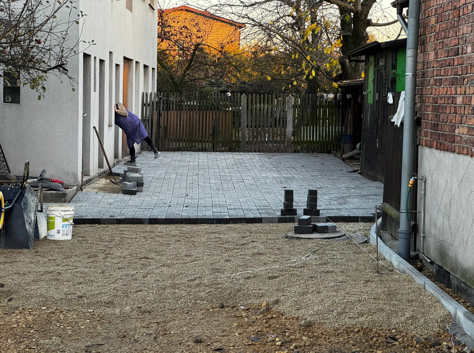 Fragment układania szarej kostki brukowej na podsypce żwirowej, widoczne narzędzia i częściowo ułożona nawierzchnia przy budynku z drewnianym ogrodzeniem w tle.