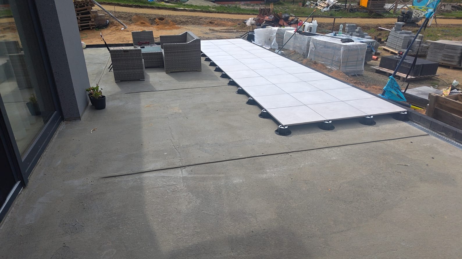 Montaż tarasu z płyt na regulowanych wspornikach. Widoczne szare płyty tarasowe, meble ogrodowe i przygotowane podłoże. Prace w toku, widok z boku.