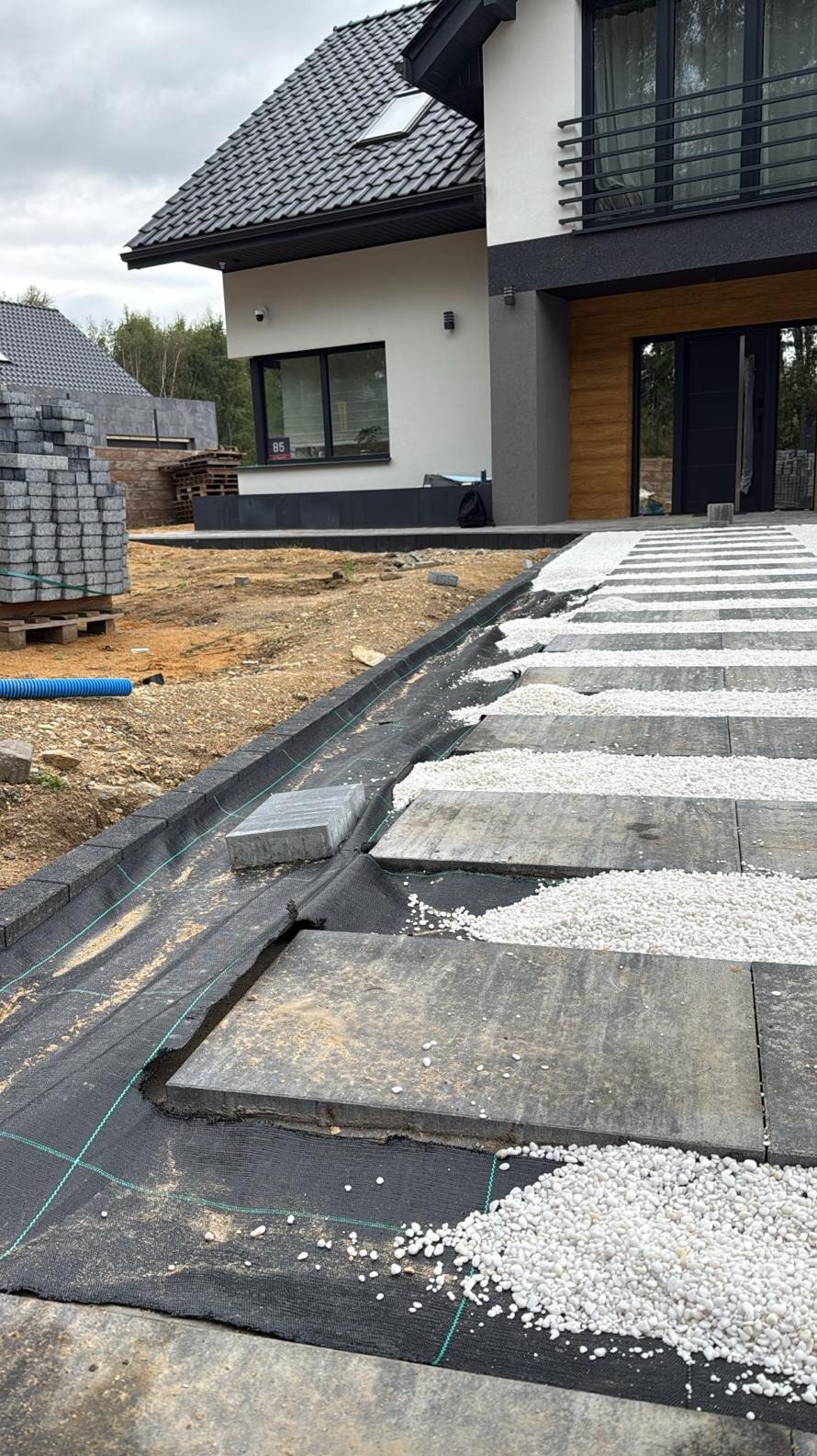 Układanie kostki: widok na częściowo ułożony chodnik z szarych płyt i białego żwiru, z widoczną warstwą geowłókniny i krawężnikiem. W tle dom z czarnym dachem.