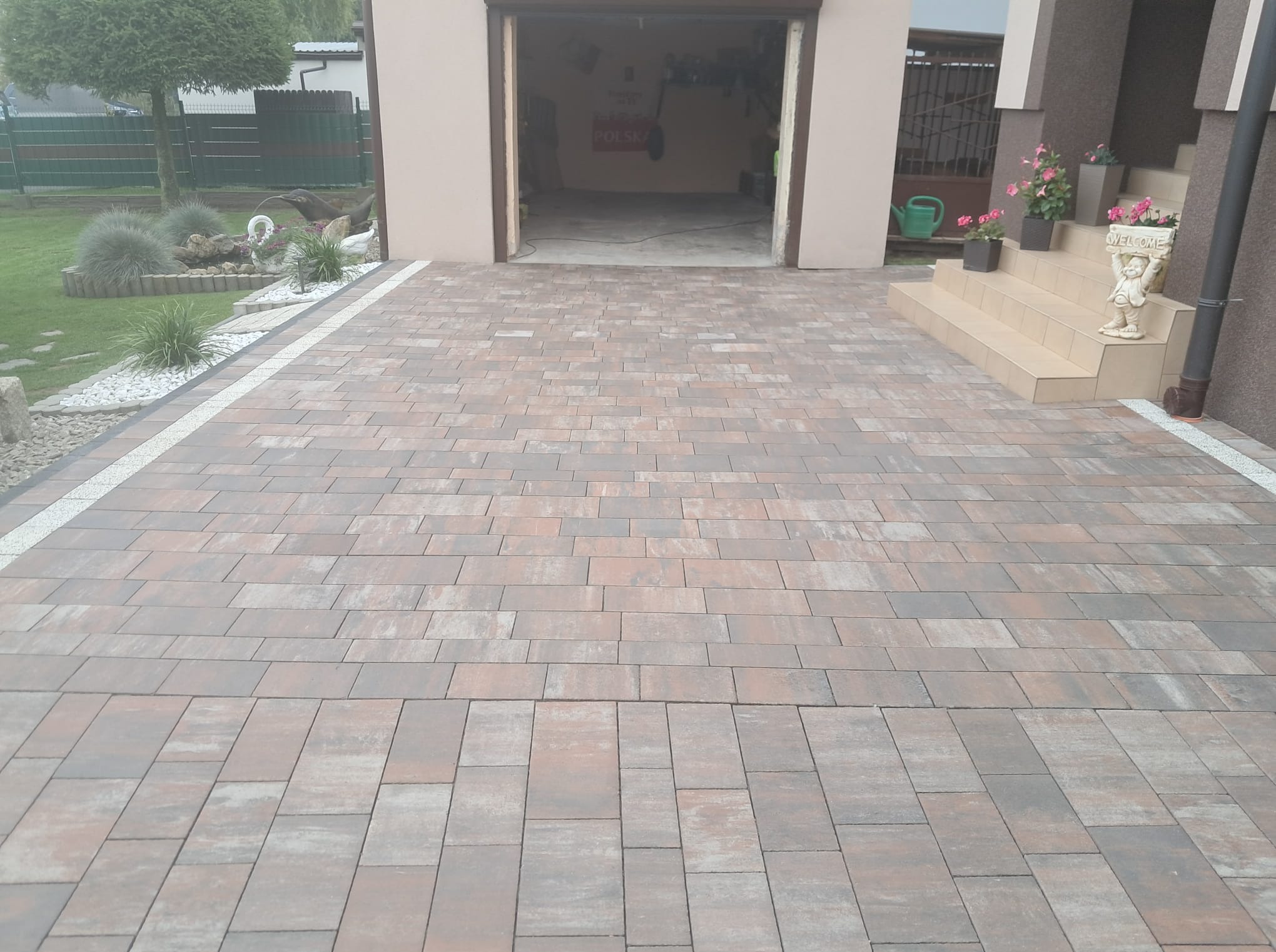 Ułożona kostka brukowa w odcieniach brązu i szarości prowadząca do garażu i schodów wejściowych. Białe obramowanie kostki. Dekoracje ogrodowe w tle.