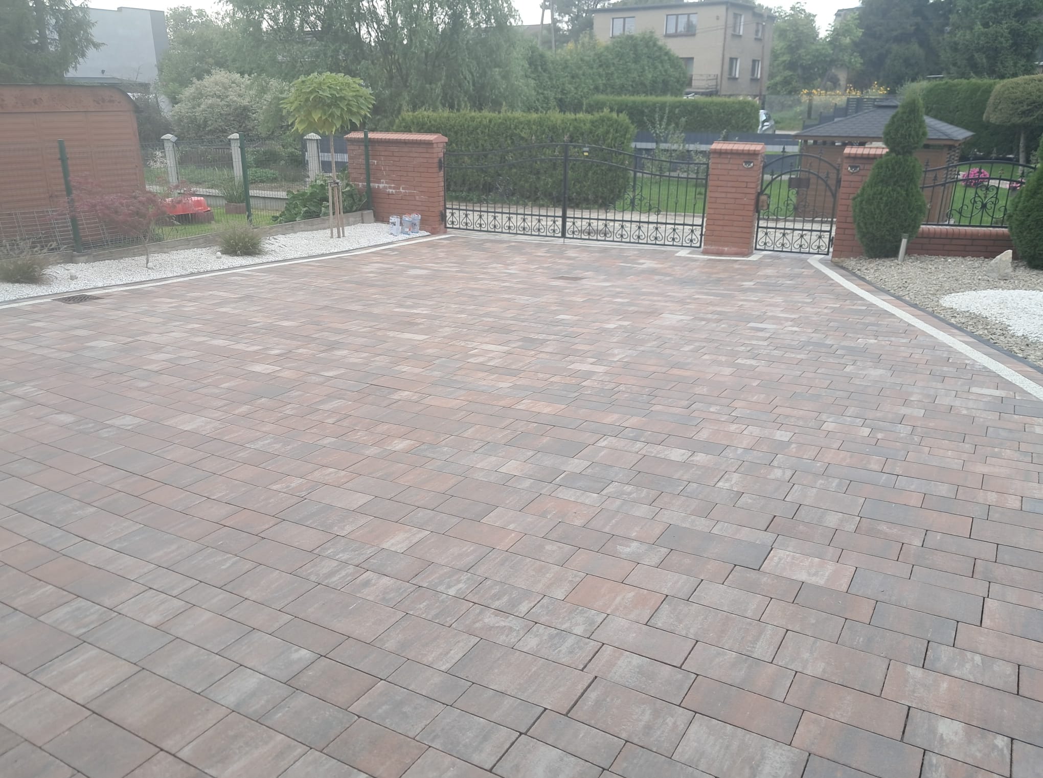Ułożona kostka brukowa w odcieniach brązu i szarości, z białymi obrzeżami, prowadząca do bramy z ceglanymi słupkami. Widoczne elementy ogrodu i zabudowa mieszkalna w tle.