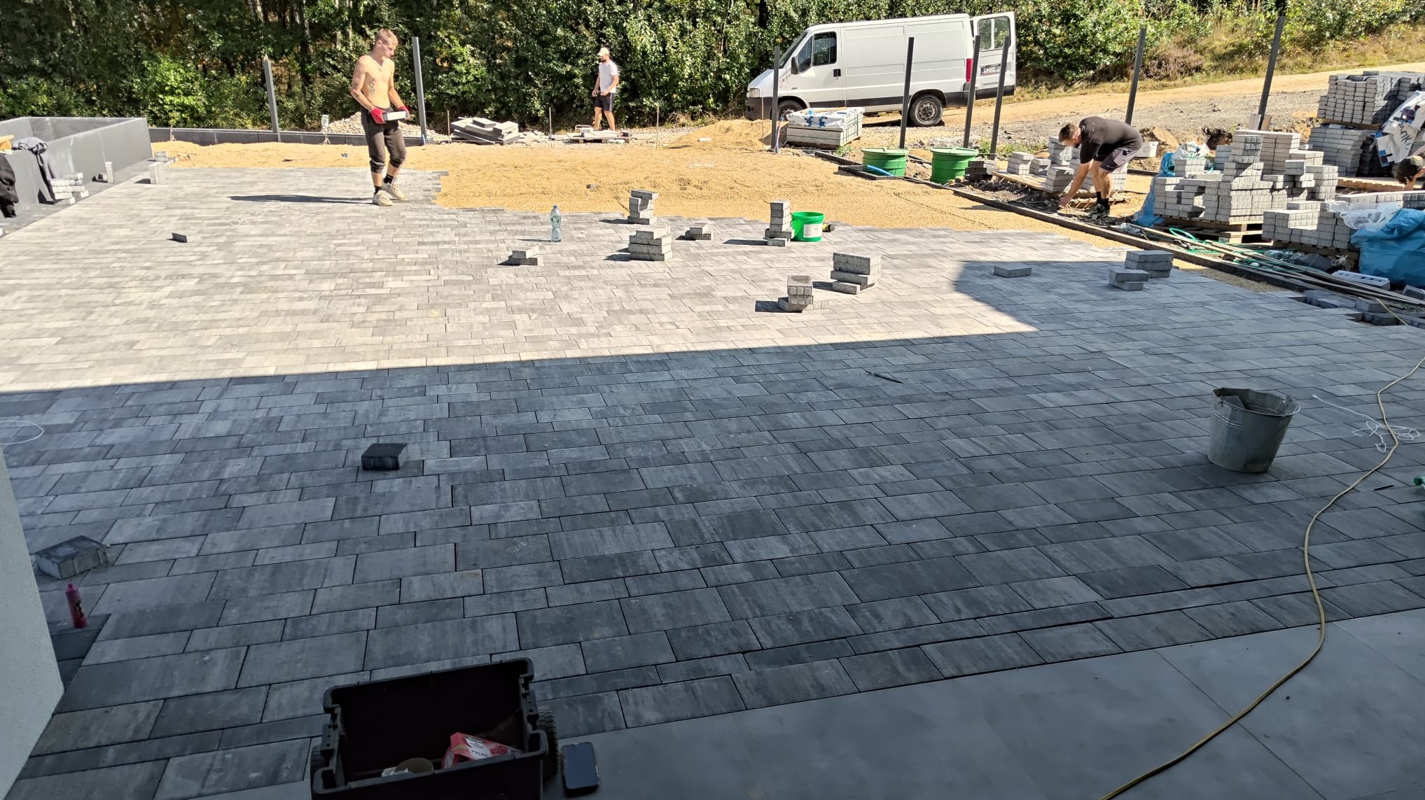 Układanie szarej kostki brukowej na posesji. Widoczni pracownicy, przygotowane materiały i częściowo ułożona nawierzchnia. Praca w toku, jasne oświetlenie.