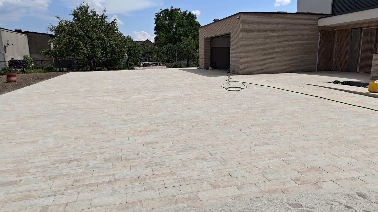 Jasna, beżowa kostka brukowa ułożona na dużej powierzchni podjazdu, z widocznym budynkiem garażowym w tle i zielonym wężem ogrodowym. Słoneczny dzień.