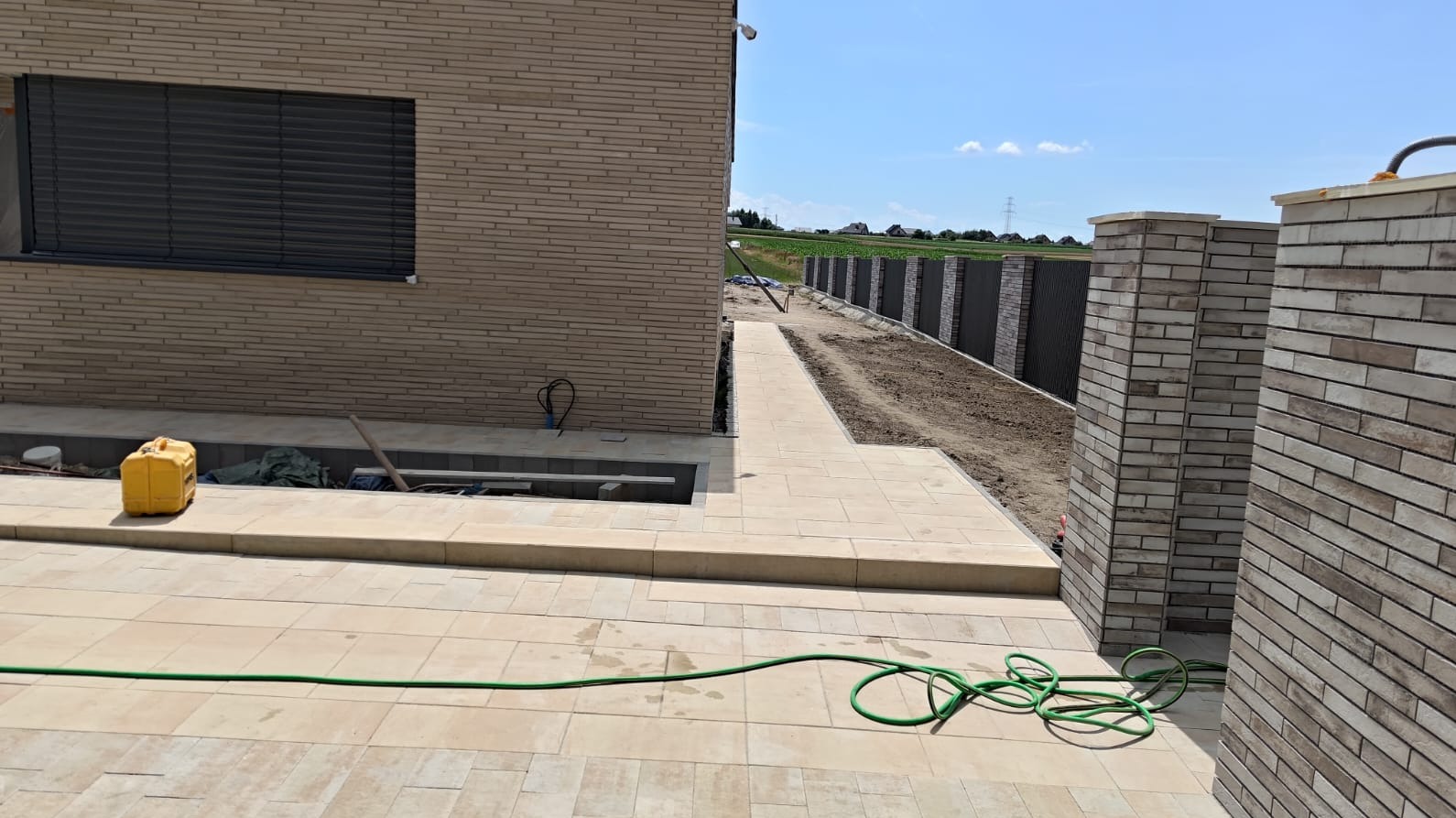 Jasna kostka brukowa tworzy ścieżkę i taras przy domu z cegły. W tle widoczne ogrodzenie panelowe i zielone pole. Wąż ogrodowy leży na kostce.