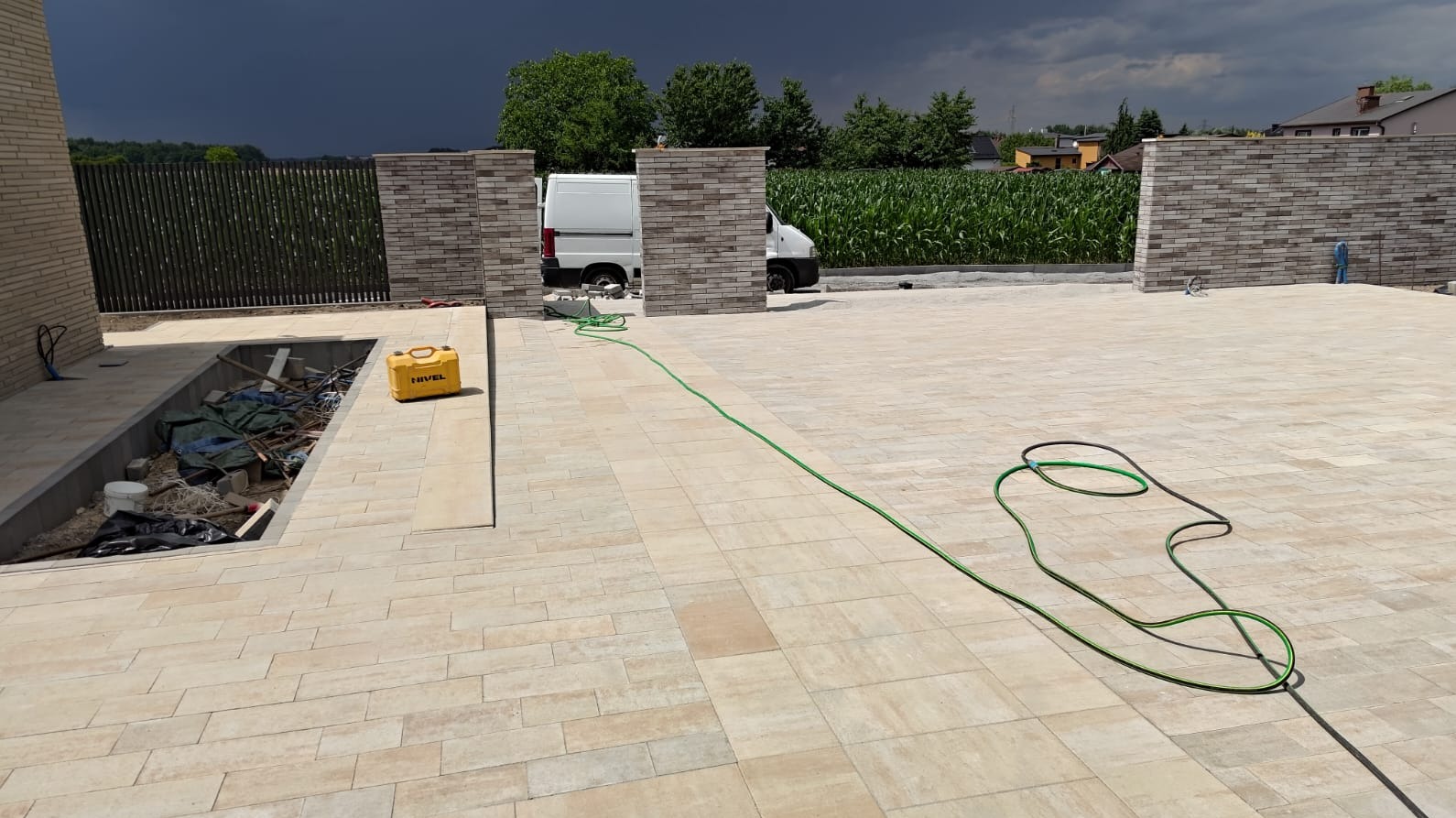 Jasna kostka brukowa na posesji z fragmentem ogrodzenia z cegły i zielonym wężem ogrodowym. W tle zaparkowany biały bus i ciemne chmury.