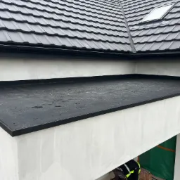 Widok z g&oacute;ry na dach z czarnej dach&oacute;wki ceramicznej i płaską powierzchnią pokrytą czarną membraną, z widocznym oknem dachowym i fragmentem elewacji budynku w trakcie wykańczania.