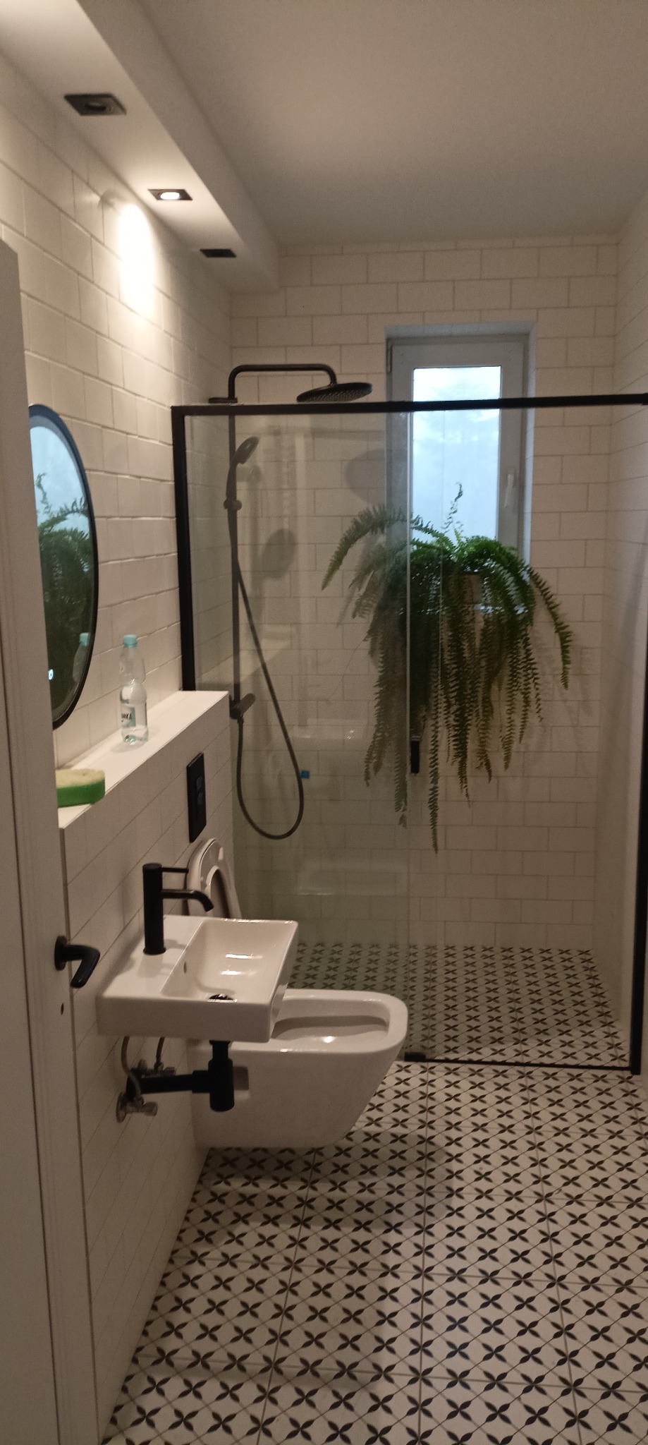 Nowoczesna łazienka z białą cegłą na ścianach, czarną armaturą i kabiną prysznicową typu walk-in, akcentem dekoracyjnym jest duża paproć w kabinie prysznicowej.