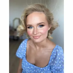 Portret uśmiechniętej blondynki z delikatnym makijażem i niebieskiej sukience w kwiatki, z widocznym odbiciem lustra w tle.